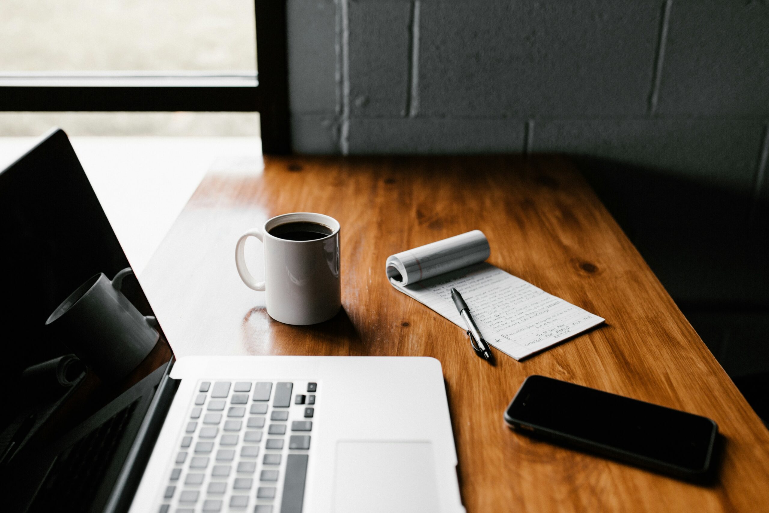 An open laptop, mug of coffee, notepad, and phone on a table.